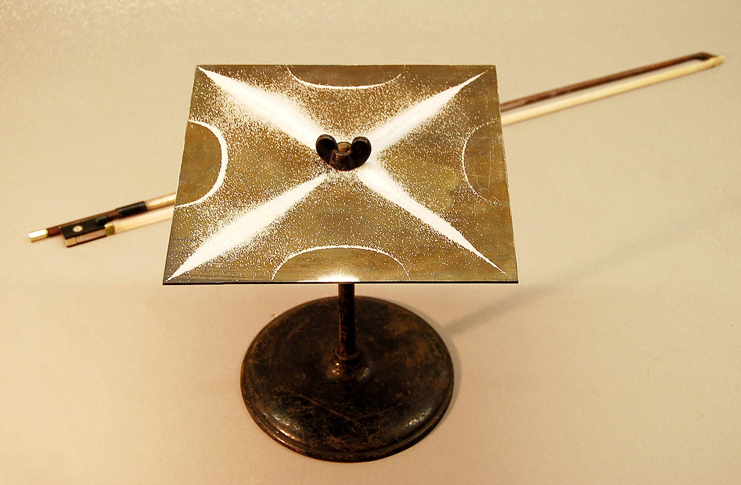 a metal Chlandi Plate has a violin bow behind it, and there is sand in the shape of a star on top