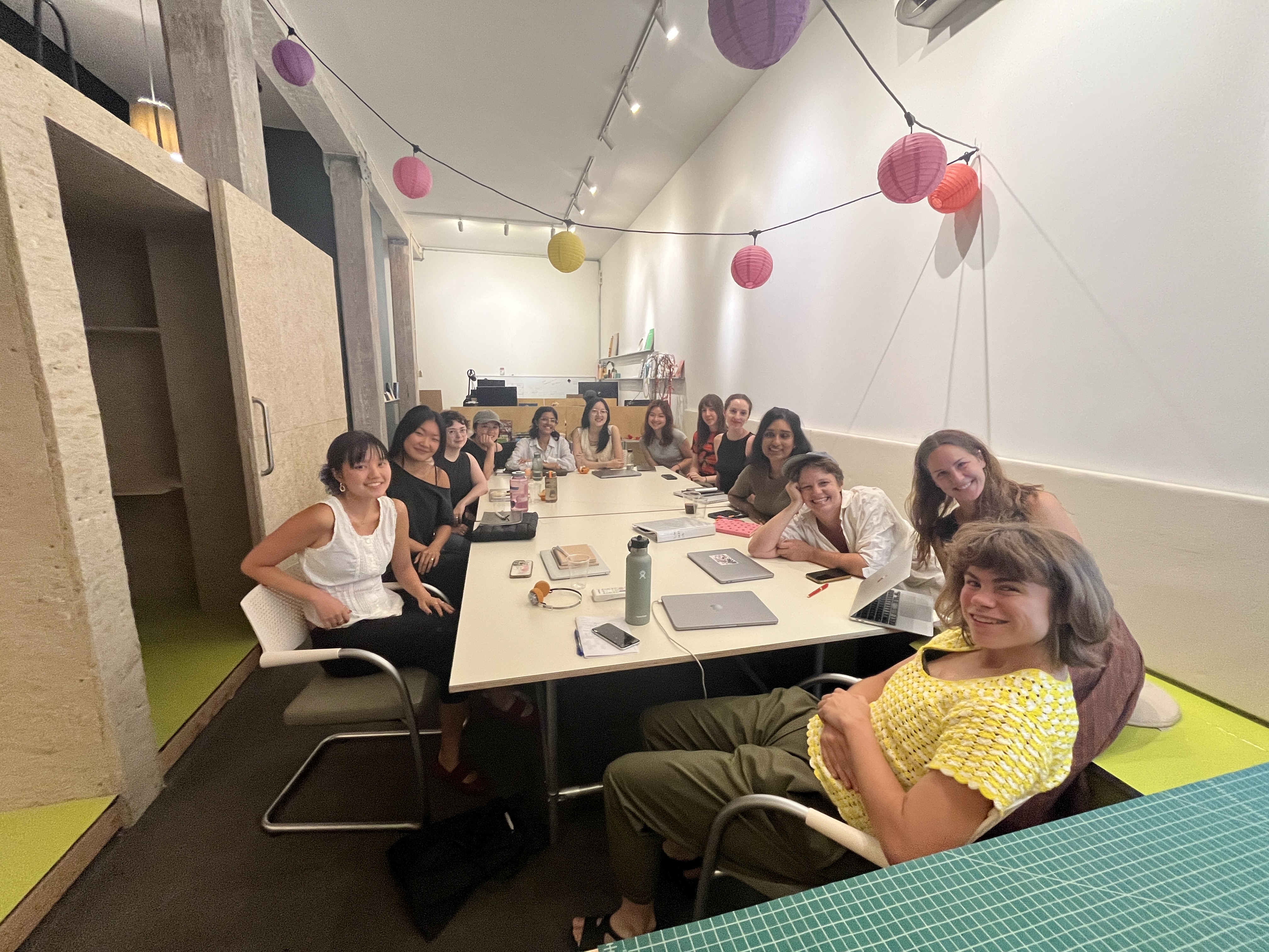 Tiff, Jill, Hannah M, Ariana, Alicia, Hannah Z, Kara, Nat, Jess, Kim, Meg, and Laurel sit around a long rectangular table smiling for the camera. Above them, pink, purple, and yellow paper lanterns are strung up, adorning the space (Dean's List) where the workshop was held.